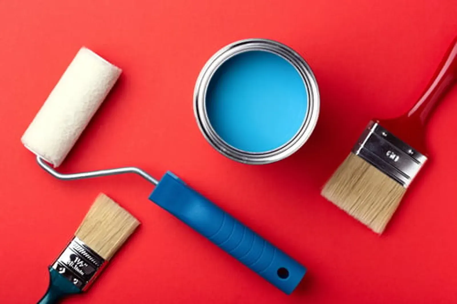 Paint roller and brush with an open can of blue paint on a bold red background